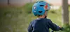 A child wearing a blue helmet with a dinosaur design is riding a bicycle outdoors. Produktbild-Vorschau