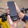 A person is holding a smartphone attached to a bike mount, with a yellow bicycle in the background. Produktbild-Vorschau