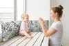A mother and baby are sitting on a wooden bench by a window. The mother is clapping her hands and smiling at the baby, who is looking at her and smiling. There are two patterned pillows behind the baby. Produktbild-Vorschau
