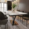 A modern dining room with a white marble-topped dining table with a black metal V-shaped base, surrounded by dark upholstered chairs. The room features a stone accent wall, dark curtains, and a large window. Voir les images du produit