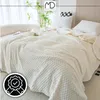 A white textured blanket is draped over a bed, with pillows and a plant in the background. Náhled obrázku produktu