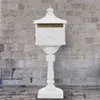 A white vintage-style mailbox on a pedestal against a brick wall. Produktbild-Vorschau