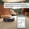 A robot vacuum cleaner is shown on a carpet next to a bed where a person and a dog are relaxing. A smartphone screen displays a map of the rooms. Náhled obrázku produktu