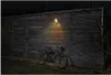 A dark gray LED floodlight is mounted on a wooden wall, illuminating a bicycle parked in front of it. Produktbild-Vorschau