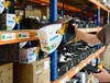 A person is holding a product box in a warehouse setting with shelves of products in the background. Produktbild-Vorschau