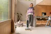 A woman is vacuuming a floor in a modern living room with a dog in a bed nearby. Produktbild-Vorschau