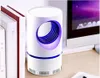 A white cylindrical insect lamp with a blue accent ring and a purple light is placed on a desk next to books and a computer monitor. Produktbild-Vorschau