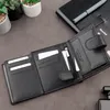 An open black leather wallet with cards and a zipper compartment is displayed on a dark gray surface, with some potted plants and a notebook in the background. Produktbild-Vorschau