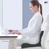 A woman is sitting at a desk and using a laptop, with an orthopedic seat cushion supporting her back. Produktbild-Vorschau