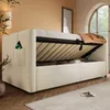 A beige upholstered bed frame with its storage compartment open, revealing the wooden slats and some stored items. A phone is visible in a side pocket. Produktbild-Vorschau