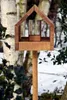 A wooden birdhouse with a glass cylinder inside, mounted on a wooden pole, in a snowy outdoor setting with trees in the background. Produktbild-Vorschau