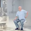 An elderly man is sitting on a foldable shower stool in a modern bathroom, holding his lower back. Voir les images du produit