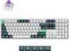 A top-down view of a white mechanical keyboard with dark green and light green keycaps, and a single purple switch shown above it. Voir les images du produit