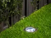 A solar ground light is embedded in artificial grass next to a dark wooden fence with hanging vines. Produktbild-Vorschau