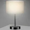 A table lamp with a white cylindrical shade and a silver metallic base and stem, placed on a white surface against a light gray background. Náhled obrázku produktu