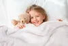 A young girl is lying in bed with a teddy bear, covered by a blanket. Produktbild-Vorschau