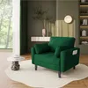 A green armchair with two pillows and two side pockets is placed on a white rug in a living room setting. The background features a window with green curtains, a shelf with decorative items, and a wall lamp. Produktbild-Vorschau