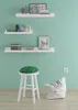 A white wooden stool with a green cushion sits on a wooden floor in front of a light green wall. There are shelves with decorative items and books on the wall, and a white beanbag chair on the floor. Produktbild-Vorschau