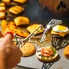 A hand using tongs to flip food on a grill, with burgers and other food items visible. Podgląd zdjęcia produktu