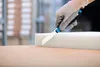 A gloved hand is using a utility knife to cut a piece of white foam on a wooden surface. Voir les images du produit
