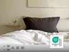 A bed with a dark pillow and white bedding, featuring laundry care symbols and a certification logo. Produktbild-Vorschau