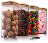 Vier klare Glasvorratsbehälter mit quadratischen Bambusdeckeln, gefüllt mit Walnüssen, Kaffeebohnen, bunten Bonbons und rosa Marshmallows, mit einem weißen Kreidestift davor Produktbild-Vorschau