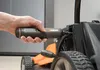 A hand is holding a black and orange electric screwdriver and is about to use it on a lawnmower. Produktbild-Vorschau