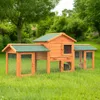 A wooden rabbit hutch with a green roof sits on a grassy lawn, surrounded by trees and bushes. Produktbild-Vorschau