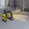 A yellow and black industrial ceramic fan heater is shown in a construction or renovation setting, with orange and yellow heat waves emanating from it. Produktbild-Vorschau