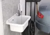 A white sink with a faucet is mounted on a gray, textured wall in a garage setting, next to a car tire and a car. Anteprima dell’immagine prodotto