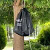 A black camping shower bag is hanging from a tree branch in a natural outdoor setting. Produktbild-Vorschau