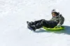 A child is sledding down a snowy slope on a green saucer sled. Produktbild-Vorschau