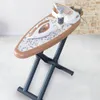 A toy ironing board set with an iron, spray bottle, and steam station on a patterned surface, set against a brick wall and tiled floor. Produktbild-Vorschau