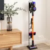 A Dyson vacuum cleaner and its accessories are neatly organized on a black stand next to a wooden cabinet with a vase of greenery. Produktbild-Vorschau