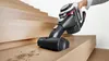 A hand holding a cordless vacuum cleaner is shown cleaning dust from wooden stairs. Produktbild-Vorschau