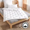 A white duvet is neatly folded on a wooden bed frame, with pillows in the background. A cotton icon is visible in the bottom right corner. Produktbild-Vorschau