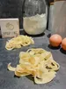 A pile of fresh pasta on a dark countertop, with a box of 'Pasta Disc' and eggs in the background. Produktbild-Vorschau