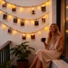 A woman sits on a ledge, holding a mug, with a string of fairy lights and photos hanging on the wall behind her. Náhled obrázku produktu
