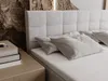 A close-up view of a bed with a light beige upholstered headboard and pillows, next to a nightstand with books. Náhled obrázku produktu