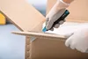 A person wearing gloves is using a utility knife to open a cardboard box. Voir les images du produit