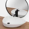 A white oval countertop sink with black faucets is shown in front of a round mirror, reflecting the sink and faucets. The sink sits on a wooden surface. Anteprima dell’immagine prodotto