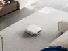 A white robot vacuum cleaner is cleaning a light gray carpet in a modern living room. Náhled obrázku produktu