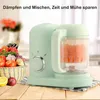 A baby food maker is shown on a wooden table with a basket of vegetables and fruits next to it. The product is mint green and has a transparent container. Produktbild-Vorschau