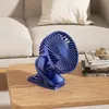 A blue clip-on fan sits on a wooden table next to a stack of books and a teacup, with a couch in the background. Produktbild-Vorschau