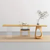 A modern dining table with a wooden top and a unique wooden base, set against a plain white wall and a light-colored rug. A tea set and a vase with branches are placed on the table. Produktbild-Vorschau