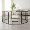 A black metal pet playpen is set up on a light-colored wooden floor in a room with a sofa and a plant in the background. Náhled obrázku produktu