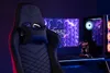 A gaming chair is shown in a dimly lit room with a computer setup. Produktbild-Vorschau