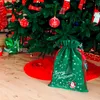 A green Christmas gift bag with 'Merry Christmas' text and Santa Claus illustration sits on a wooden floor under a decorated Christmas tree. Produktbild-Vorschau
