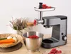 A kitchen machine with a meat grinder attachment is shown in use, grinding meat into a bowl. Vegetables are visible in a basket nearby. Podgląd zdjęcia produktu