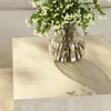 A close-up shot of a glass vase filled with white flowers and green leaves, placed on a beige surface with dappled sunlight casting shadows. Produktbild-Vorschau
